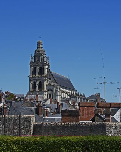 A large stone cathedral with a prominent central tower and dome, viewed from a distance over rooftops and a stone wall under a clear blue sky.
