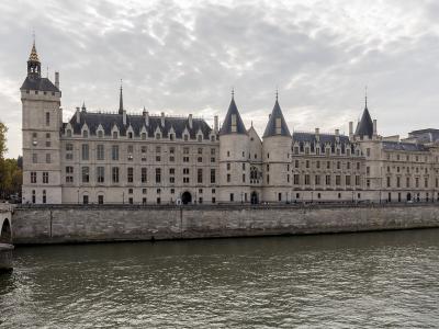 The image shows a long, historic stone building with conical-roofed towers and arched windows situated along the bank of a wide river, with a tour boat on the water and a bridge in the distance under a partly cloudy sky.