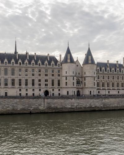The image shows a long, historic stone building with conical-roofed towers and arched windows situated along the bank of a wide river, with a tour boat on the water and a bridge in the distance under a partly cloudy sky.