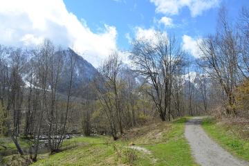 1. A gravel hiking trail winds through a grassy, wooded area with bare trees and a small stream on the left, leading toward snow-dusted mountains under a blue sky with clouds.
