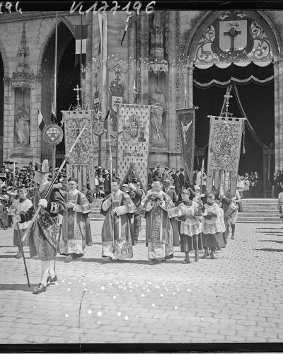 A black and white photograph showing a religious procession exiting the Gothic facade of a cathedral, with participants carrying banners and flags, including one reading "Jésus Regina," and a crowd watching from the sides.