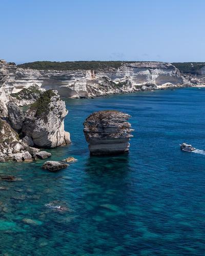 A panoramic view of the white limestone cliffs of Bonifacio, Corsica, featuring a prominent sea stack (the "Perruque") and a winding path along the cliff edge, with boats on the blue Mediterranean Sea.