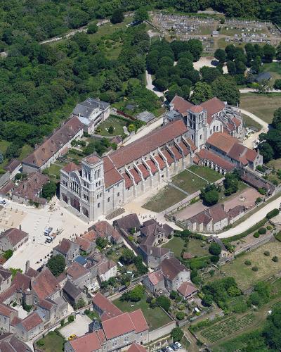 An aerial photograph showing the Basilique Sainte-Marie-Madeleine de Vézelay, a large Romanesque abbey church with a prominent facade and towers, surrounded by the historic town of Vézelay, a cemetery, and vineyards.