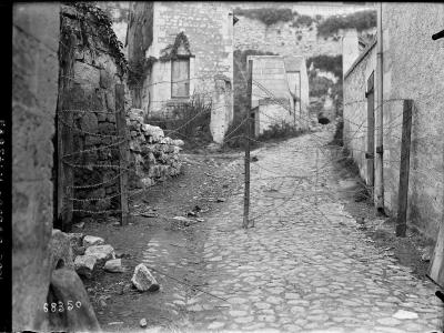A black and white historical photograph showing a cobblestone path blocked by barbed wire strung between wooden posts, with stone buildings and rubble visible in the background.