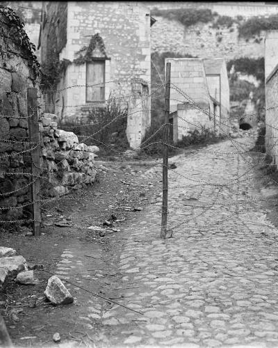 A black and white historical photograph showing a cobblestone path blocked by barbed wire strung between wooden posts, with stone buildings and rubble visible in the background.