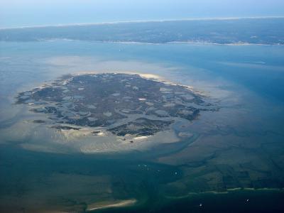 An aerial photograph showing a small, irregularly shaped island surrounded by shallow blue water and tidal flats, with a distant coastline visible in the background; no text or signage is present.