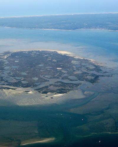 An aerial photograph showing a small, irregularly shaped island surrounded by shallow blue water and tidal flats, with a distant coastline visible in the background; no text or signage is present.