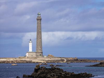 A tall, slender stone lighthouse stands on a rocky coastal island next to a smaller white lighthouse, with a small boat moored in the foreground water under a cloudy sky.
