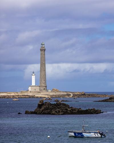 A tall, slender stone lighthouse stands on a rocky coastal island next to a smaller white lighthouse, with a small boat moored in the foreground water under a cloudy sky.