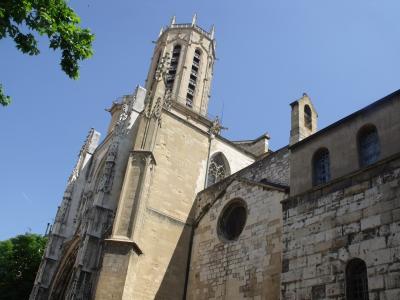 A low-angle view of a historic stone cathedral featuring a prominent Gothic bell tower with pinnacles, adjacent to older Romanesque-style walls with round and arched windows, under a clear blue sky with tree foliage in the corner.