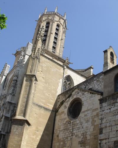 A low-angle view of a historic stone cathedral featuring a prominent Gothic bell tower with pinnacles, adjacent to older Romanesque-style walls with round and arched windows, under a clear blue sky with tree foliage in the corner.