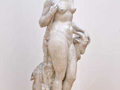 A full-length white marble sculpture of a standing nude female figure holding a goat’s head, displayed on a wooden platform inside a gallery with light wood flooring and white walls.