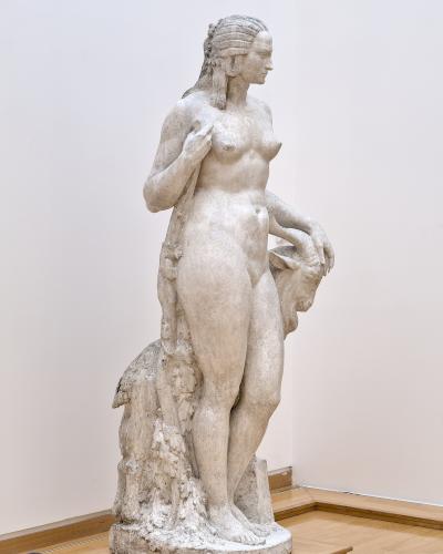 A full-length white marble sculpture of a standing nude female figure holding a goat’s head, displayed on a wooden platform inside a gallery with light wood flooring and white walls.