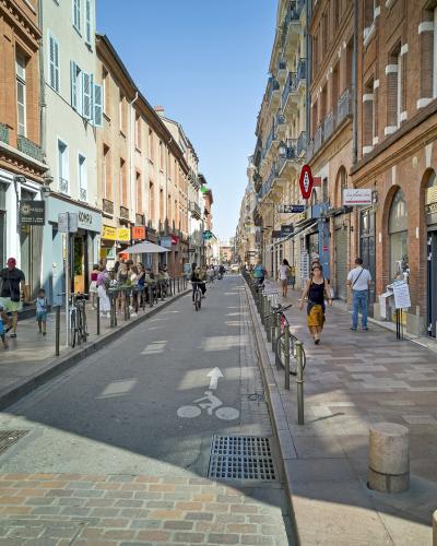 A narrow, sunlit pedestrian and bicycle street lined with multi-story brick buildings featuring arched ground-floor arcades and shuttered windows, with signage for "Bio Bon" and a bus stop visible on the left.