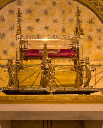 The image shows a golden reliquary shrine containing a relic wrapped in red cloth, set against a vaulted ceiling painted with stars and geometric patterns, with three golden statues flanking the base.