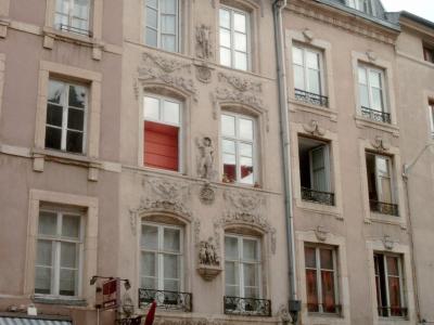 A multi-story building with ornate stone carvings and sculptures on its facade, located on a street with shops like "Stylos Cave à" and "PRINCESSE tam-tam" at the ground level.