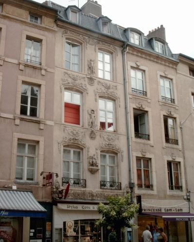 A multi-story building with ornate stone carvings and sculptures on its facade, located on a street with shops like "Stylos Cave à" and "PRINCESSE tam-tam" at the ground level.