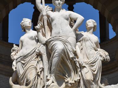 A large white marble fountain sculpture featuring a central female figure holding a staff, flanked by two other female figures and two bull heads at the base from which water flows, set within an arched stone structure.