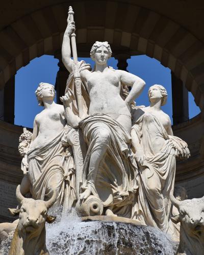 A large white marble fountain sculpture featuring a central female figure holding a staff, flanked by two other female figures and two bull heads at the base from which water flows, set within an arched stone structure.