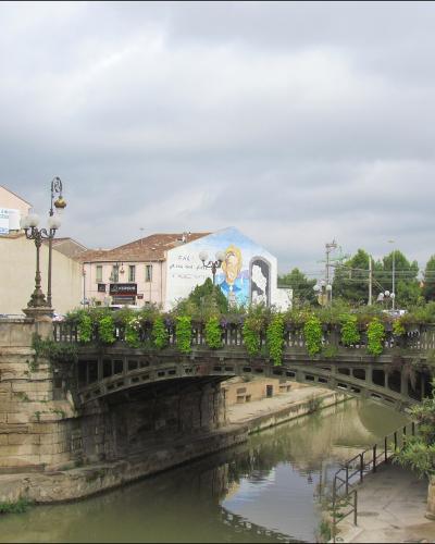 A stone bridge with ornate iron railings and hanging flower boxes spans a narrow canal in an urban setting, with buildings featuring French signage (e.g., "E.Leclerc", "VERSACE") and a mural visible in the background under an overcast sky.
