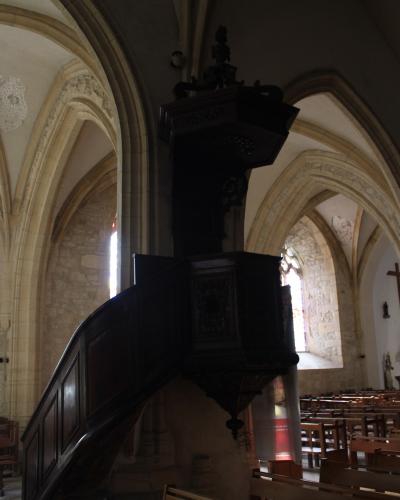 The image shows the interior of a church featuring a prominent, dark wooden pulpit with a curved staircase, set against stone walls and Gothic-style vaulted arches.