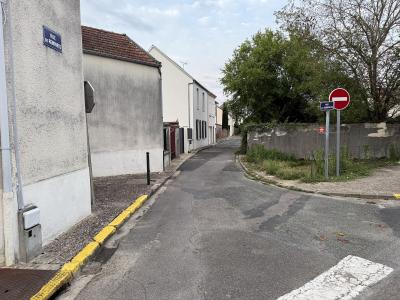 A narrow paved street in a French town with a blue street sign on the left wall reading "RUE DES REMPARTS" and a matching sign on a pole to the right, alongside a "No Entry" traffic sign and a concrete wall bordering a green area.