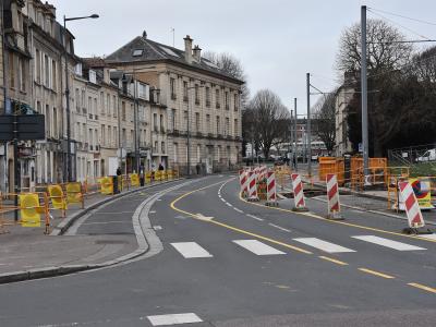 A street scene showing roadworks with yellow and red-and-white striped barriers on a curved urban road lined with multi-story stone buildings under an overcast sky.