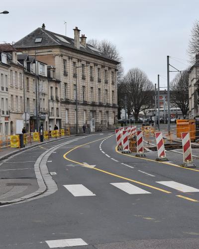 A street scene showing roadworks with yellow and red-and-white striped barriers on a curved urban road lined with multi-story stone buildings under an overcast sky.
