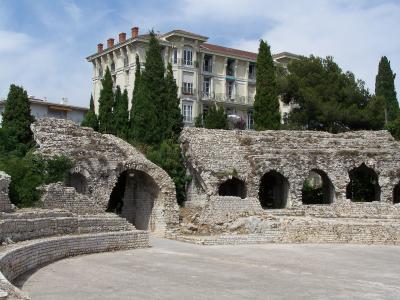 The image shows ancient stone ruins with arched openings and tiered seating, set against a backdrop of a large multi-story residential building and tall cypress trees under a partly cloudy sky.