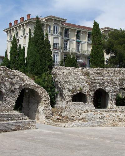 The image shows ancient stone ruins with arched openings and tiered seating, set against a backdrop of a large multi-story residential building and tall cypress trees under a partly cloudy sky.