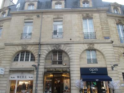 A street-level view of a classical stone building facade featuring three ground-floor storefronts (WEILL, Mac Douglas, apm MONACO) and upper floors with arched windows and wrought-iron balconies.