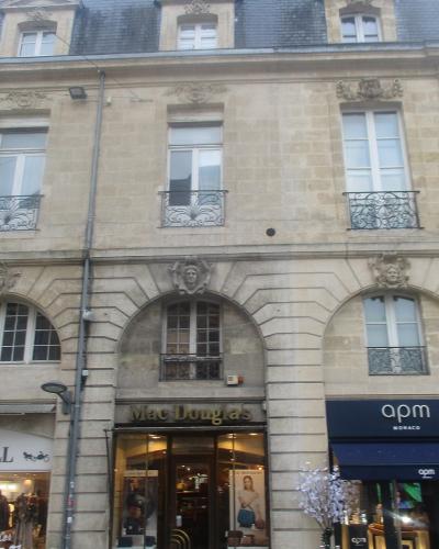 A street-level view of a classical stone building facade featuring three ground-floor storefronts (WEILL, Mac Douglas, apm MONACO) and upper floors with arched windows and wrought-iron balconies.
