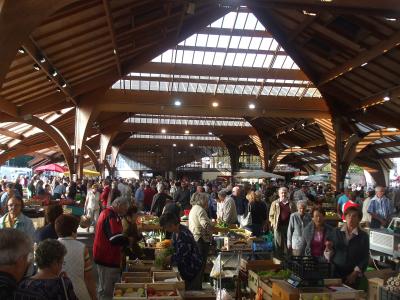 A crowded indoor market hall with distinctive wooden arched trusses and skylights, filled with shoppers and stalls selling fresh produce like fruits and vegetables.