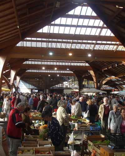 A crowded indoor market hall with distinctive wooden arched trusses and skylights, filled with shoppers and stalls selling fresh produce like fruits and vegetables.