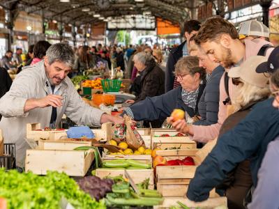 The image shows a crowded indoor market stall with vendors and customers interacting over wooden crates of fresh produce like lemons, peppers, and lettuce under a high, industrial-style metal roof.