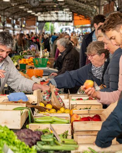 The image shows a crowded indoor market stall with vendors and customers interacting over wooden crates of fresh produce like lemons, peppers, and lettuce under a high, industrial-style metal roof.