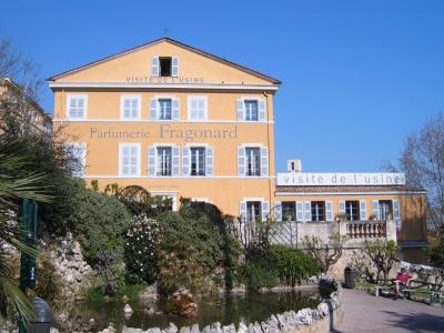 A yellow 18th-century building with white shutters, featuring large text on the facade reading "Parfumerie Fragonard" and "VISITE DE L'USINE", set against a blue sky with a garden and pond in the foreground.