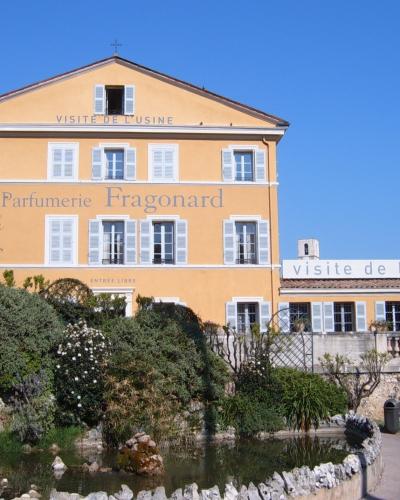 A yellow 18th-century building with white shutters, featuring large text on the facade reading "Parfumerie Fragonard" and "VISITE DE L'USINE", set against a blue sky with a garden and pond in the foreground.