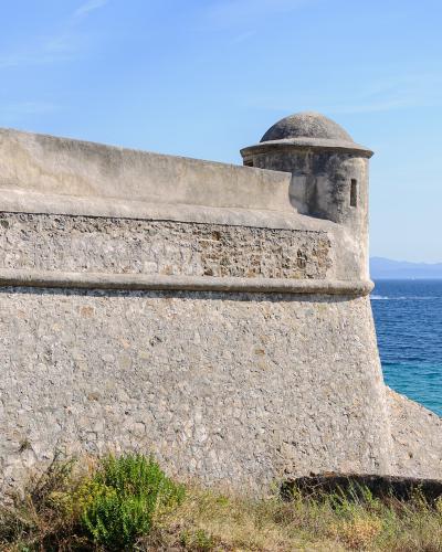 Stone fortress wall and watchtower overlooking the blue sea under a clear sky.