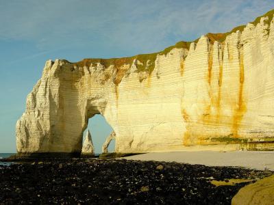 The image shows a large natural chalk arch (La Manneporte) and a sea stack (Aval) at the base of towering white cliffs along a pebble beach, with the English Channel in the background under a partly cloudy sky.