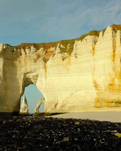 The image shows a large natural chalk arch (La Manneporte) and a sea stack (Aval) at the base of towering white cliffs along a pebble beach, with the English Channel in the background under a partly cloudy sky.