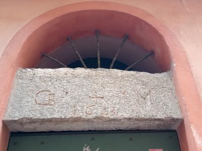 A close-up of a stone door lintel with the carved initials "C B" and "M" flanking a cross and the date "1623", set within a pink stucco archway above a green door.