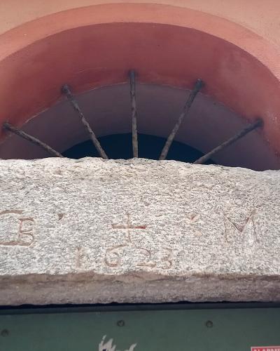 A close-up of a stone door lintel with the carved initials "C B" and "M" flanking a cross and the date "1623", set within a pink stucco archway above a green door.
