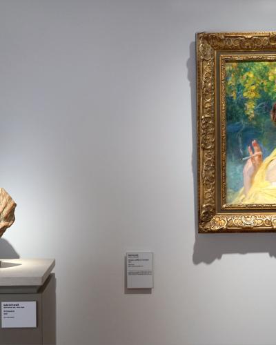 The image shows an interior museum gallery with a terracotta bust on a pedestal and a framed oil painting of a woman hanging on the wall, with small informational placards visible.