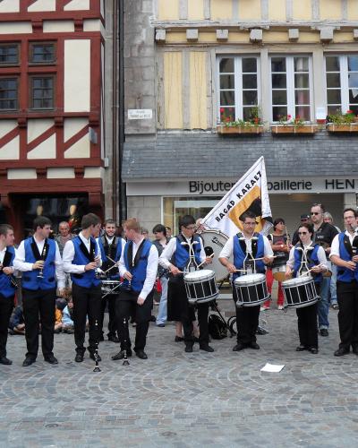 A bagad (Breton traditional music band) in blue and white uniforms performs on a cobblestone street in front of half-timbered buildings, with a banner reading "BAGAD KARAEZ" and a shop sign for "Bijouterie HENTIC".