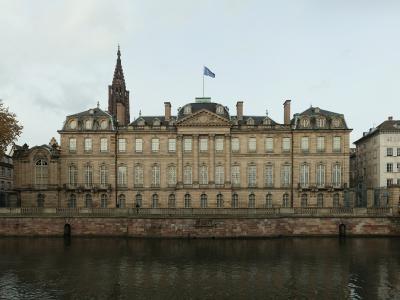 A wide-angle view of the Palais Rohan in Strasbourg, France, showing its grand Baroque facade along the Ill river, with the spire of Strasbourg Cathedral visible behind it and a blue flag flying atop the building.