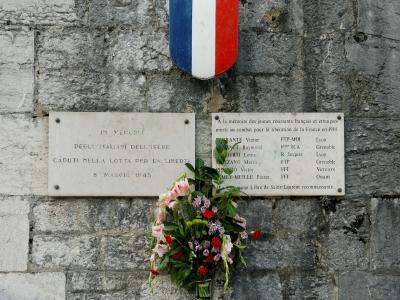 A stone wall featuring two memorial plaques and a French tricolor shield, with one plaque in Italian honoring Italians of Isère and another in French listing names of young French and foreign resistance fighters who died in 1944, signed by “La Commun