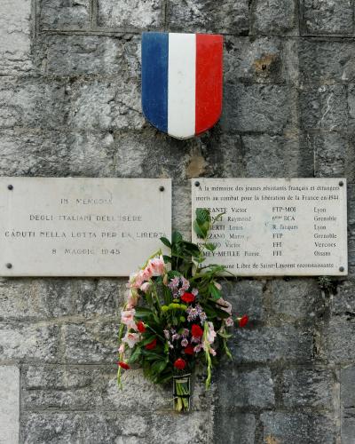 A stone wall featuring two memorial plaques and a French tricolor shield, with one plaque in Italian honoring Italians of Isère and another in French listing names of young French and foreign resistance fighters who died in 1944, signed by “La Commun