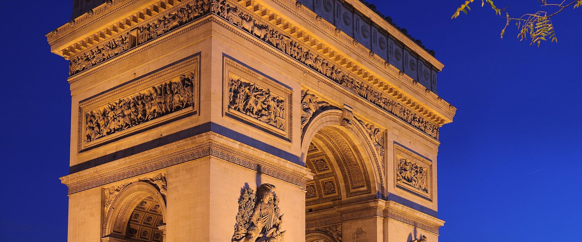 The image shows the Arc de Triomphe in Paris, France, illuminated at night with light trails from traffic and the Eiffel Tower visible in the background.