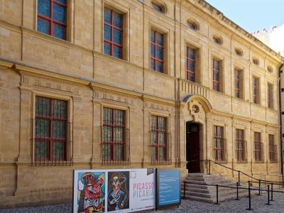 The image shows the exterior of a classical stone building with red-framed windows, barred lower windows, and a central arched entrance; signage in front advertises a "PICASSO PICABIA" exhibition at "GRANET," indicating it is the Musée Granet in A